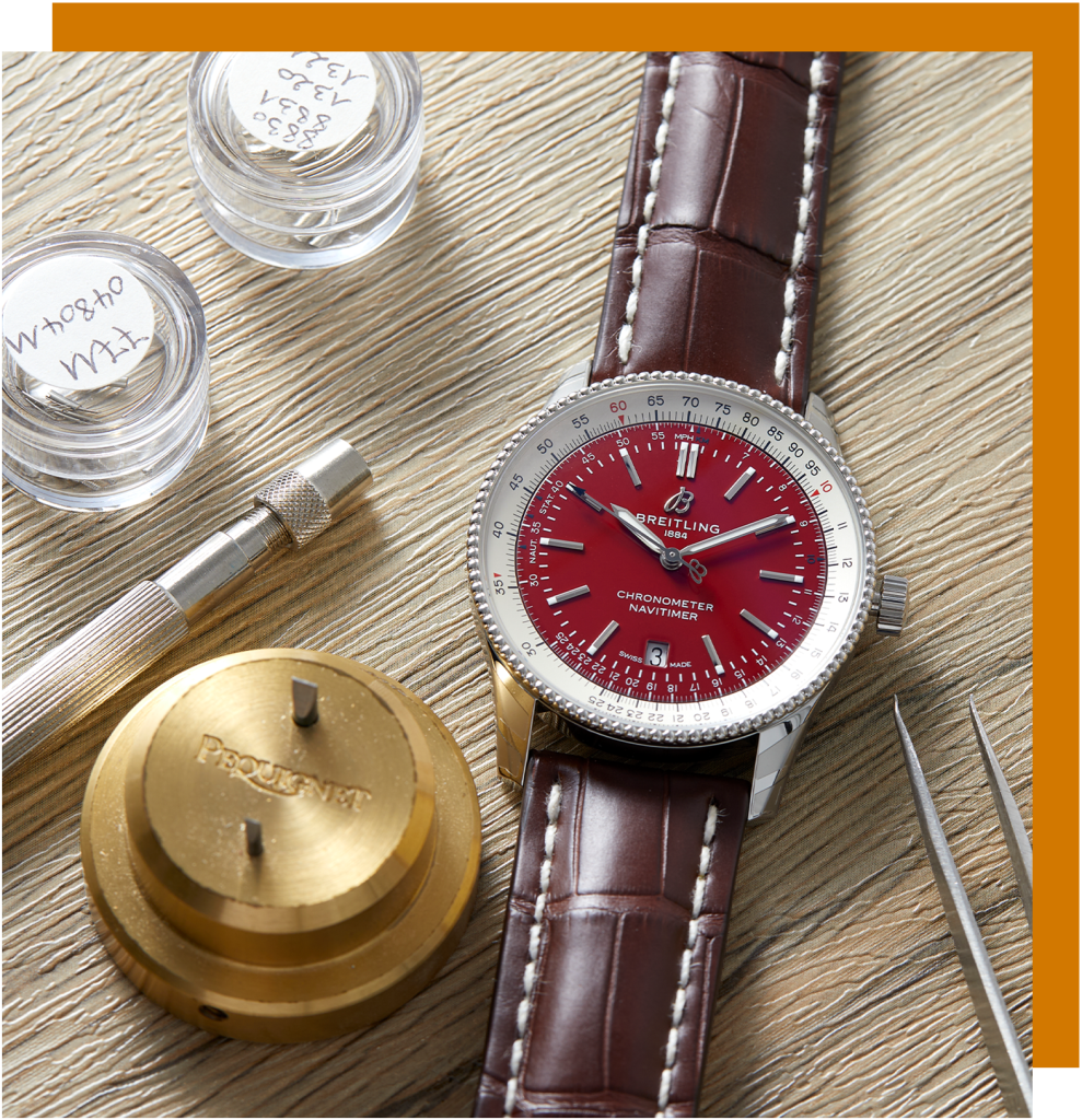 Luxury Breitling Navitimer watch with a red dial, brown leather strap, and accompanying watchmaking tools on a wooden surface.